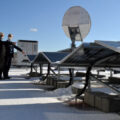 町役場庁舎屋上の太陽光パネル前に立つ小松町長（左）。冬季は雪の反射を取り入れられるよう両面に発電パネルを備える。奥に立つのは蓄電池